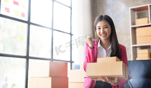 自由职业亚洲女性的初创 SME 小企业企业家使用带盒子的笔记本电脑快乐成功快乐的亚洲女性她的手举起在线营销包装和交付 SME 理念