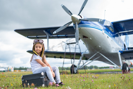 一个穿着飞行员服装的小女孩坐在一个手提箱上，背景是一架带螺旋桨的飞机。