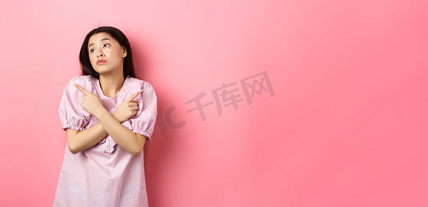 优柔寡断的亚洲女性无法决定，手指横着，看起来沉思，做出选择，站在粉红色背景中困惑