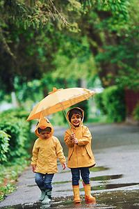 两个带雨伞的孩子穿着黄色防水斗篷和靴子在雨后一起在户外玩耍