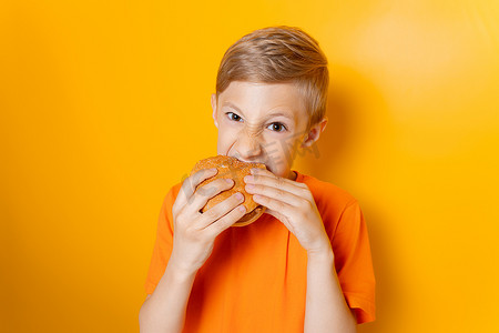 一个穿橙色 T 恤的男孩双手拿着一个汉堡包，在黄色背景下贪婪地咬着它