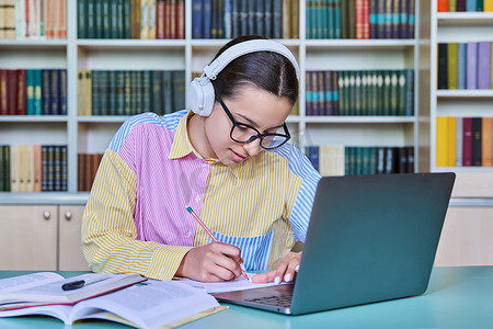 耳机桌摄影照片_十几岁的女学生在图书馆学习，使用笔记本电脑