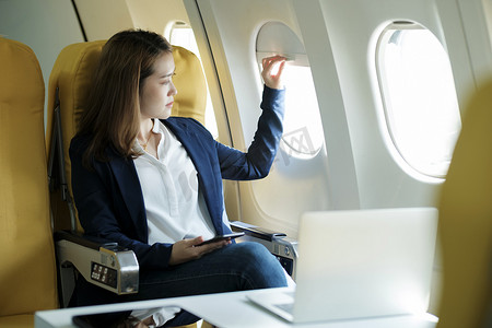 女商人在飞机上，使用笔记本电脑和电话工作，看看飞机窗口。