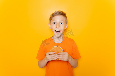 一个穿着橙色 T 恤的快乐男孩站着，手里拿着一个汉堡包，对着镜头微笑