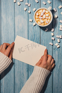 女手拿着空的问候语或邀请卡，在木制蓝色背景上用白杯咖啡和棉花糖进行模拟。