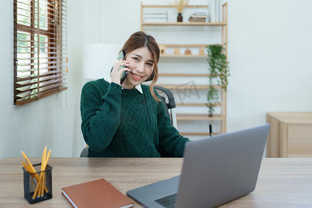 一位亚洲年轻女性的肖像，她在清晨使用办公桌上的电话、电脑和财务文件时面带微笑