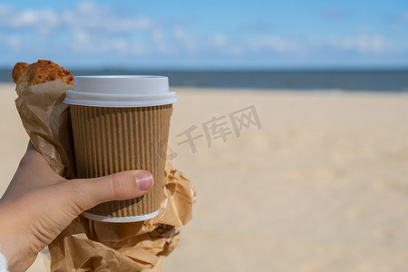 美味的新鲜羊角面包，在海滩大海背景下，女性手中的纸杯中装有咖啡。
