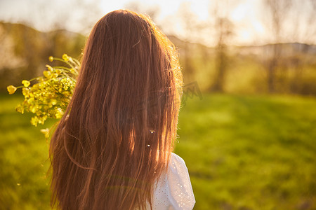 从一位红发女子的背后拍摄的照片，在夕阳的光线下拿着一束鲜花