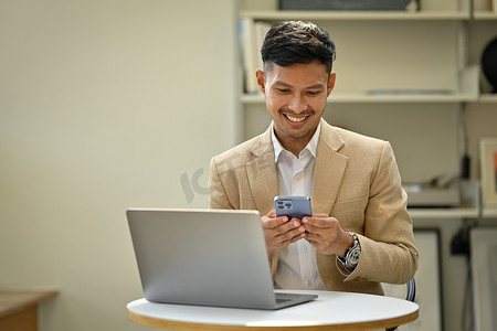 穿着时尚西装、在现代办公空间使用笔记本电脑和使用智能手机的微笑商人