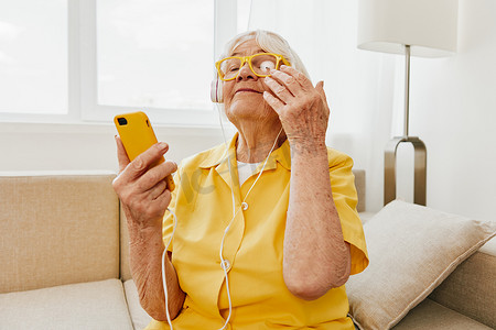 快乐的老妇人在手机上观看视频通话，在耳机上听音乐，明亮的现代室内装饰，生活方式在线交流。