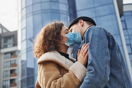 一对戴着防护面具的夫妇在检疫时间在商业大楼附近的城市里互相亲吻。