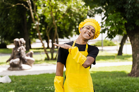 黑人厨师摄影照片_身着厨师帽和黄色围裙制服的快乐黑人非洲少年厨师在户外微笑。