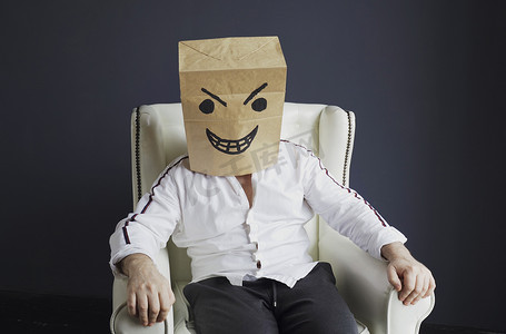 一个头上顶着纸袋的男人坐在一张白色的椅子上，脸上带着愤怒的笑脸。