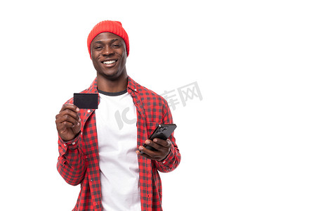 身着红色衬衫和帽子的 30 多岁英俊的深色皮肤美国男子在白色工作室背景和复制空间中为网上购物做卡片和智能手机广告