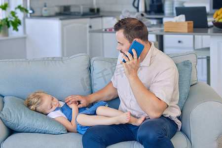 生病的小女孩躺在沙发上，爸爸用手机打电话给医生。