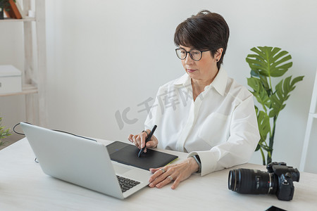 中年女性平面设计师或摄影师在现代办公室工作时使用数字图形平板电脑，专业女性修饰师坐在现代工作区，通用设计平板电脑和附近的相机