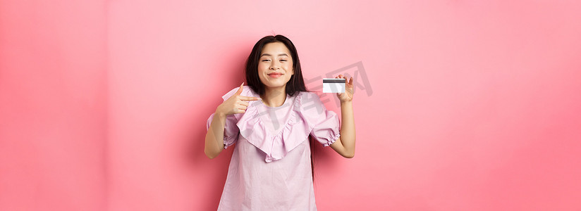 开朗可爱的亚洲女性满意地微笑着，展示塑料信用卡，穿着粉色背景的裙子