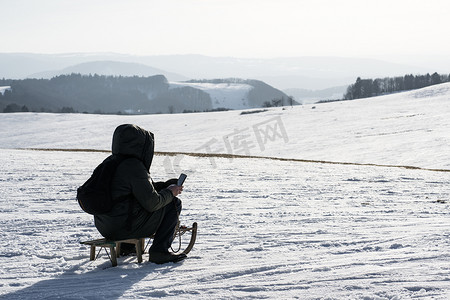 下雪保暖摄影照片_一个男人看着他的手机，冬天坐在雪山顶上的寒冷中，坐在儿童雪橇上，裹着他的冬季保暖夹克