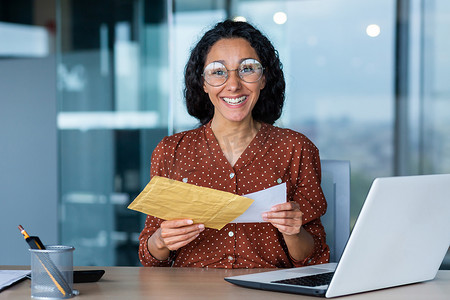 快乐的西班牙裔女性微笑着看着相机，女商人收到带有好消息通知信的信封，上班族拿着文件和笔记本电脑在办公室工作