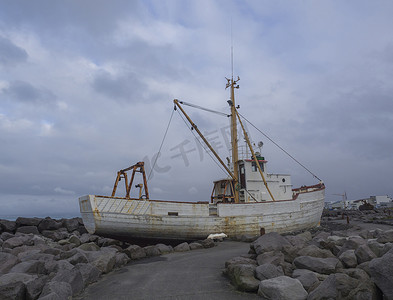 凯夫拉维克摄影照片_旧生锈的废弃渔船，站在岩石上的沉船，冰岛凯夫拉维克的海滨长廊，蓝天云背景