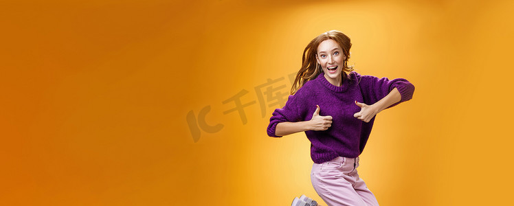穿着紫色毛衣的兴奋而快乐的风趣年轻红发女性从幸福和满足中跳出来，竖起大拇指表示赞同，给予积极答复，喜欢很棒的衣服