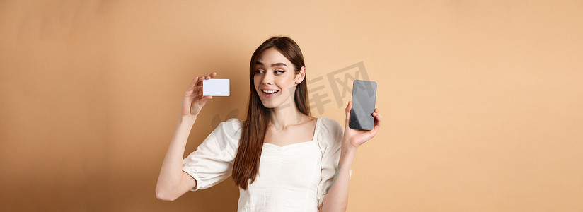 站在米色背景上，年轻的白人女性带着高兴的微笑展示塑料信用卡，展示空的手机屏幕