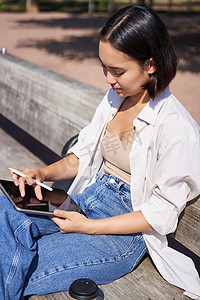 图形创意画摄影照片_年轻的亚洲创意女孩用笔在平板电脑上画画，坐在公园的户外，画画