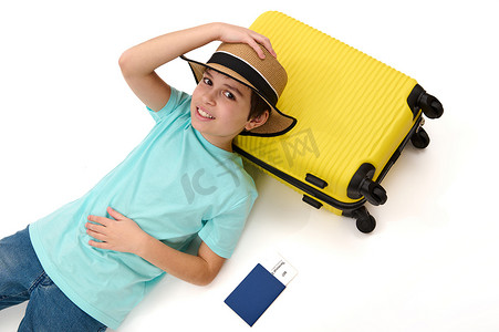 行李箱男孩摄影照片_穿着休闲服和草帽的轻松青春期前男孩躺在黄色手提箱上，去国外度暑假