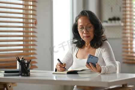 中年女士在手机上阅读信息并在笔记本上做笔记的肖像