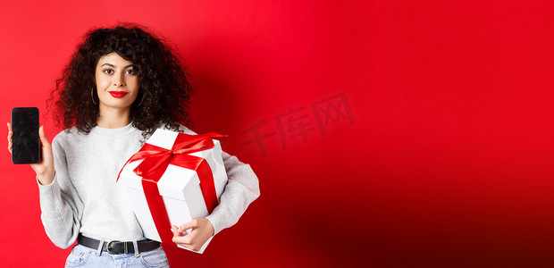 购物app摄影照片_一头卷发的美女，在空荡荡的智能手机屏幕上展示购物应用程序，拿着包裹在节日盒子里的礼物，站在红色背景上