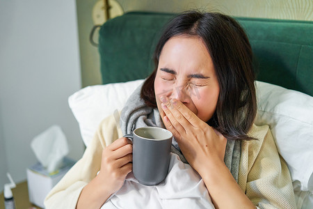 亚洲女孩在床上的特写，感觉不舒服，感冒打喷嚏，擦流鼻涕，喝热茶