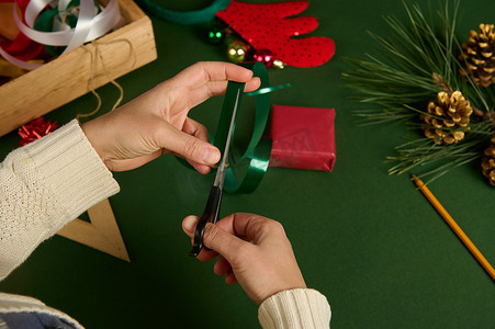 赛事时间摄影照片_一位女士将闪亮的绿色胶带切成条状，用于装饰用红色包装纸包裹的圣诞礼物的顶视图