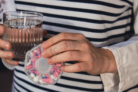 女手分拣药片组织者每周拍摄一杯水特写药盒，里面装有药片，每天服用白色粉红色药物和胶囊。