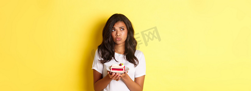 优柔寡断、悲伤的非洲裔美国女性站在黄色背景上，看起来心烦意乱，不能吃蛋糕