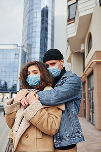 感染摄影照片_在检疫时间，一对戴着防护面具的夫妇在商业大楼附近的城市里互相拥抱。