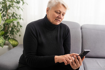 快乐成熟的 60 多岁老妇人拿着智能手机使用手机应用程序进行视频通话，一边看有趣的视频一边笑，坐在家里的沙发上兴奋地在手机上赢得在线彩票竞标