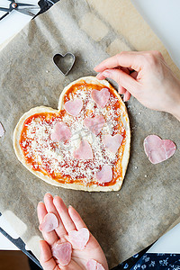 心形披萨面团，厨师在披萨上涂上番茄酱，撒上帕玛森芝士，并用火腿装饰成心形。