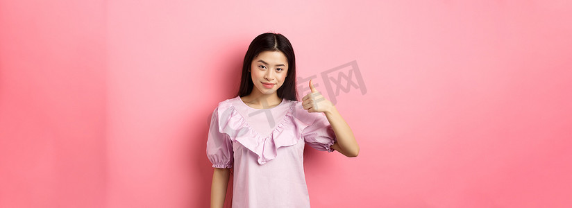 微笑的亚洲女孩竖起大拇指表示赞同，称赞不错的选择，喜欢并推荐产品，站在粉红色背景中