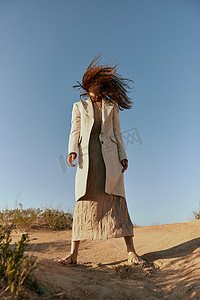 一位身穿时尚夹克和长裙的女士站在沙滩上，背景是晴朗的天空，她的头发在运动中遮住了脸