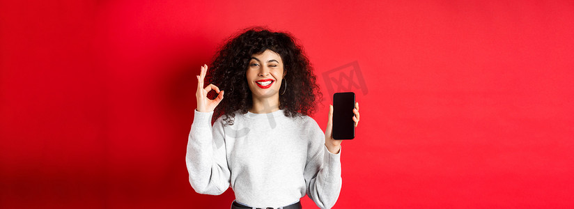 购物app摄影照片_有智能手机的迷人女性，显示好的标志和空的电话屏幕，推荐购物应用程序，站在红色背景下