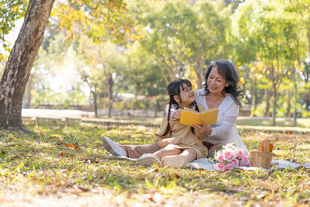 手绘讲故事摄影照片_快乐的亚洲祖母讲故事，周末一起在美丽的公园野餐时，给她可爱的孙女读故事。