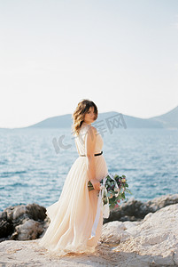 拿着花束的新娘侧身站在多岩石的海边