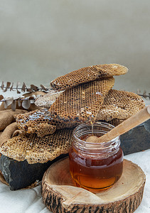 美味的蜂蜜摄影照片_美味的蜂蜜从玻璃瓶上的新鲜蜂窝中滴落，黑色岩石上装有木制蜂蜜勺。