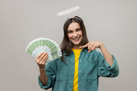 一堆钞票摄影照片_头上有光环的女人指着一堆欧元纸币，微笑着获利。