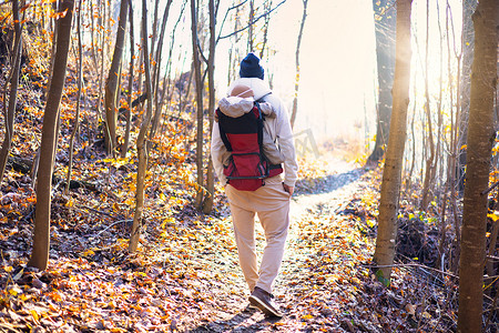 运动型父亲背着他的小儿子穿着冬季连身衣和帽子，背着背包在秋天的森林里徒步旅行。