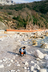 捡摄影照片_爸爸蹲在一个小女孩旁边，在青山背景下，从水边的岩石海滩捡起鹅卵石