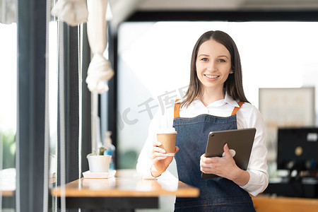 开一家小企业，一个穿着围裙的快乐亚洲女人站在酒吧柜台咖啡店附近，小企业主、餐厅、咖啡师、咖啡馆、在线、中小企业、企业家和卖家概念