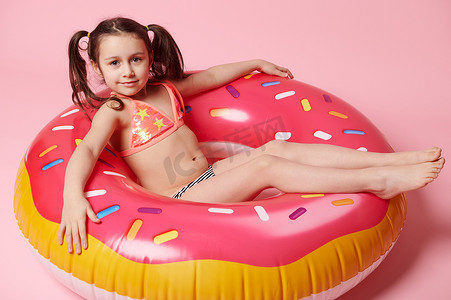 从上方看迷人的小女孩在漂浮的甜甜圈游泳圈上放松，与粉色背景隔离