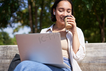 亚洲女孩坐在笔记本电脑和无线耳机旁，喝咖啡，看屏幕，做作业，远程工作