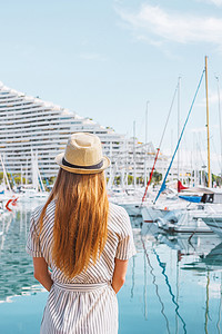 戴着帽子的金发女人背对着大海、游艇和小船背景站着。
